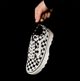A hand holding a black and white checkerboard patterned sneaker against a solid black background. The sneaker features a thick white sole and white laces, with a classic lace-up design. The minimalist setting emphasizes the shoe's bold pattern.
