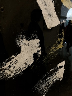 A dark, abstract image featuring broad, textured white paint strokes against a black background. There is a faint silhouette of a person visible, creating an element of mystery.
