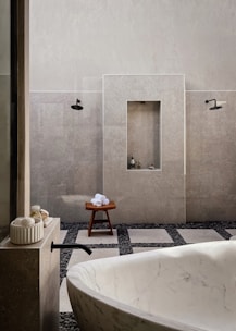 A minimalist bathroom featuring a large white marble bathtub in the foreground. The walls are tiled in a light gray stone, complemented by black fixtures including a faucet and showerheads. There is a wooden stool with rolled white towels placed on top, and small bottles are nestled in a recessed wall shelf. The floor is a mix of large square tiles and dark pebbles, adding to the spa-like ambiance.