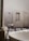 A minimalist bathroom featuring a large white marble bathtub in the foreground. The walls are tiled in a light gray stone, complemented by black fixtures including a faucet and showerheads. There is a wooden stool with rolled white towels placed on top, and small bottles are nestled in a recessed wall shelf. The floor is a mix of large square tiles and dark pebbles, adding to the spa-like ambiance.