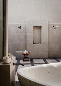 A minimalist bathroom featuring a large white marble bathtub in the foreground. The walls are tiled in a light gray stone, complemented by black fixtures including a faucet and showerheads. There is a wooden stool with rolled white towels placed on top, and small bottles are nestled in a recessed wall shelf. The floor is a mix of large square tiles and dark pebbles, adding to the spa-like ambiance.