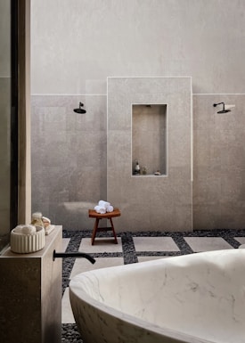 A minimalist bathroom featuring a large white marble bathtub in the foreground. The walls are tiled in a light gray stone, complemented by black fixtures including a faucet and showerheads. There is a wooden stool with rolled white towels placed on top, and small bottles are nestled in a recessed wall shelf. The floor is a mix of large square tiles and dark pebbles, adding to the spa-like ambiance.