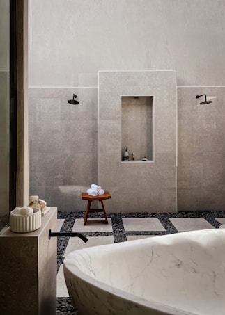 A minimalist bathroom featuring a large white marble bathtub in the foreground. The walls are tiled in a light gray stone, complemented by black fixtures including a faucet and showerheads. There is a wooden stool with rolled white towels placed on top, and small bottles are nestled in a recessed wall shelf. The floor is a mix of large square tiles and dark pebbles, adding to the spa-like ambiance.