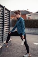 A person in athletic wear performs a stretching exercise outdoors. The individual is dressed in a blue jacket and black leggings, with one leg propped on a low wall and holding a water bottle. The background features a fence and blurred residential buildings.