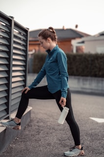 A person in athletic wear performs a stretching exercise outdoors. The individual is dressed in a blue jacket and black leggings, with one leg propped on a low wall and holding a water bottle. The background features a fence and blurred residential buildings.