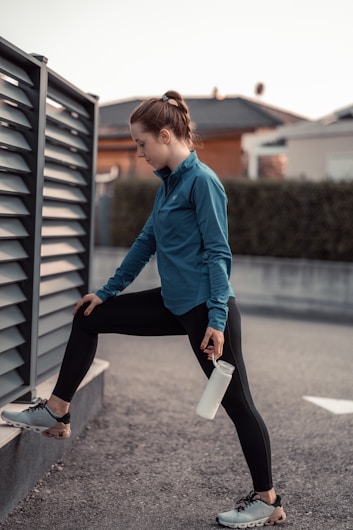 A person in athletic wear performs a stretching exercise outdoors. The individual is dressed in a blue jacket and black leggings, with one leg propped on a low wall and holding a water bottle. The background features a fence and blurred residential buildings.