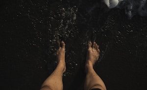 Bare feet standing on dark wet sand with small waves approaching. The image conveys a sense of calm and grounding, focusing on the connection with nature.