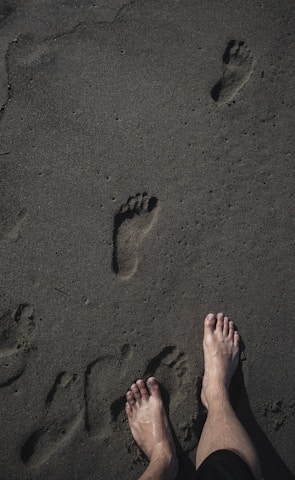 Bare feet standing on wet sand with several footprints leading away, capturing a moment by the beach or shoreline.