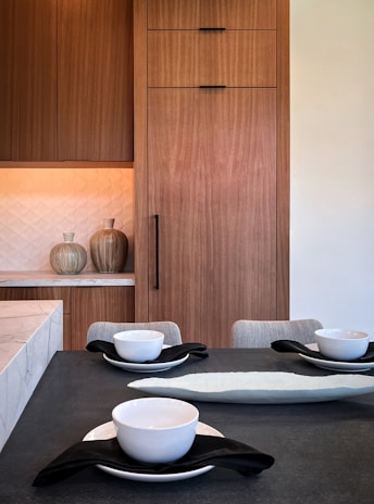 A modern kitchen or dining space featuring wooden cabinetry with clean lines. The countertop is adorned with white bowls placed on black napkins, along with a long, decorative platter. The background includes textured wall tiles and decorative vases, adding a sophisticated touch.