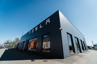 A modern, dark gray building with large windows and the word 'TESLA' displayed prominently on its side. The structure is situated on a paved area with a clear, blue sky overhead and a few trees visible in the background.