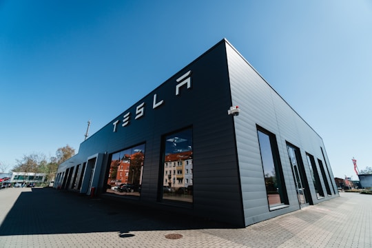 A modern, dark gray building with large windows and the word 'TESLA' displayed prominently on its side. The structure is situated on a paved area with a clear, blue sky overhead and a few trees visible in the background.