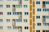 A facade of a residential building with multiple rectangular windows. Some windows have balconies bordered by bright orange frames. The structure features a repetitive pattern in a minimalist architectural style, with closed windows having white blinds or curtains.