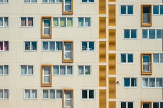 A facade of a residential building with multiple rectangular windows. Some windows have balconies bordered by bright orange frames. The structure features a repetitive pattern in a minimalist architectural style, with closed windows having white blinds or curtains.