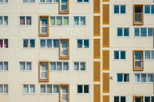 A facade of a residential building with multiple rectangular windows. Some windows have balconies bordered by bright orange frames. The structure features a repetitive pattern in a minimalist architectural style, with closed windows having white blinds or curtains.