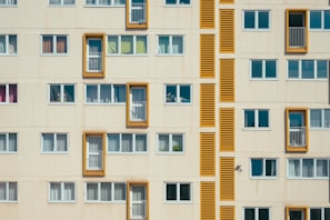 A facade of a residential building with multiple rectangular windows. Some windows have balconies bordered by bright orange frames. The structure features a repetitive pattern in a minimalist architectural style, with closed windows having white blinds or curtains.