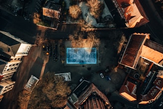 An aerial view of an urban area with multiple buildings surrounding a central blue basketball court. The sun casts long shadows from the structures and trees, giving the scene a dramatic effect. The streets and sidewalks curve around the buildings, and some leafless trees are visible, suggesting the season might be late fall or winter.