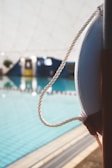 A close-up view of a white life ring attached to a vertical pole by a rope, positioned next to a swimming pool with clear blue water. In the background, an indoor setting is indicated by blurred walls and windows, suggesting a swimming facility.