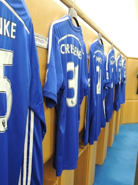 A row of blue football jerseys hangs on wooden lockers, each bearing names and numbers on the back. The setting appears to be a team's changing room with a polished, organized look.