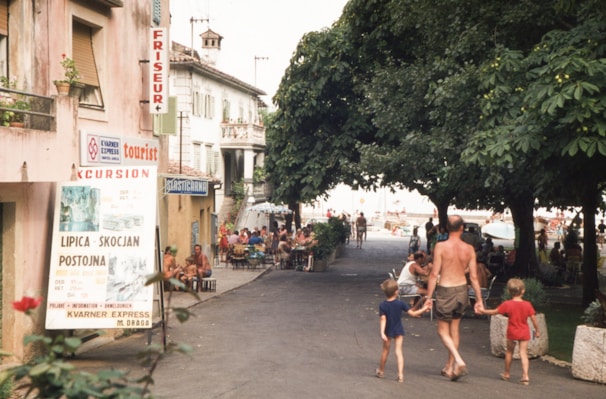 A street scene with people walking, some in swimwear, indicating a casual, leisure atmosphere. The street is lined with trees providing shade, and there are establishments such as a hairdresser and cafes with outdoor seating. A large sign promotes excursions to tourist destinations. The presence of families suggests a vacation setting.
