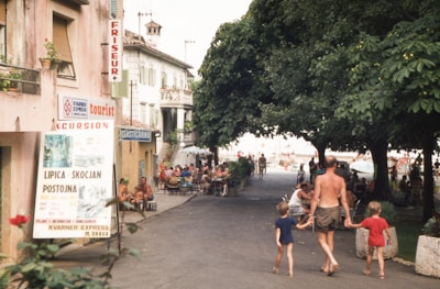 A street scene with people walking, some in swimwear, indicating a casual, leisure atmosphere. The street is lined with trees providing shade, and there are establishments such as a hairdresser and cafes with outdoor seating. A large sign promotes excursions to tourist destinations. The presence of families suggests a vacation setting.