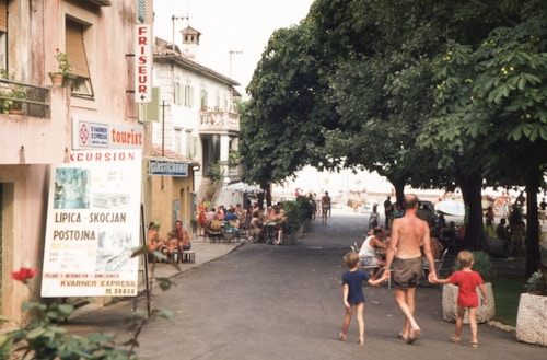 A street scene with people walking, some in swimwear, indicating a casual, leisure atmosphere. The street is lined with trees providing shade, and there are establishments such as a hairdresser and cafes with outdoor seating. A large sign promotes excursions to tourist destinations. The presence of families suggests a vacation setting.