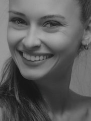 A black and white close-up portrait of a person smiling warmly, with a focus on the face and expression. The subject has long hair and is wearing an earring, with an overall positive and engaging demeanor.