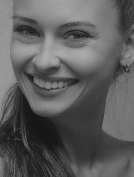 A black and white close-up portrait of a person smiling warmly, with a focus on the face and expression. The subject has long hair and is wearing an earring, with an overall positive and engaging demeanor.