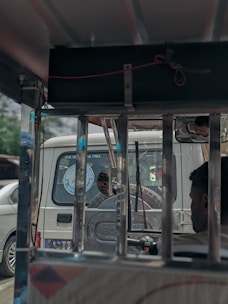 Image shows a partially visible passenger inside a vehicle, seen through a window with metal bars, looking outward. A car with a spare tire mounted on the back can be seen behind the main focus vehicle. The words 'LIVE YOUR LIFE FREE' are visible on the rear window of the car. There are also some other vehicles in the background.