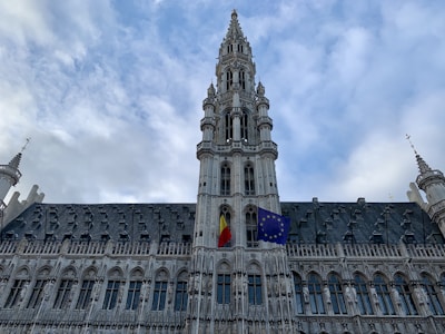 A large Gothic-style building with intricate architectural details and a tall central tower topped with spires. Two flags are prominently displayed in the center of the building, the Belgian national flag and the European Union flag. The sky is partly cloudy, providing a dramatic backdrop.