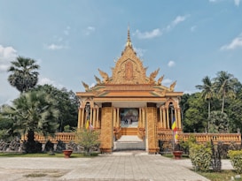 An ornate, golden temple stands majestically amidst lush greenery. The architectural design features intricate carvings and a central spire pointing towards the sky. Palm trees border the scene, complementing the peaceful, tropical setting. Flags and landscaped plants enhance the entrance, contributing to an inviting atmosphere.