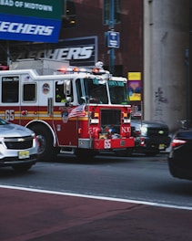 A bright red fire truck with flashing lights is driving down a busy city street. The vehicle is equipped with American flags and detailed with '65' on its side. Surrounding the truck, there are other vehicles, including a silver sedan and a larger black vehicle. The background features a muted cityscape with billboards and street signs.