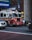 A bright red fire truck with flashing lights is driving down a busy city street. The vehicle is equipped with American flags and detailed with '65' on its side. Surrounding the truck, there are other vehicles, including a silver sedan and a larger black vehicle. The background features a muted cityscape with billboards and street signs.