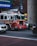 A bright red fire truck with flashing lights is driving down a busy city street. The vehicle is equipped with American flags and detailed with '65' on its side. Surrounding the truck, there are other vehicles, including a silver sedan and a larger black vehicle. The background features a muted cityscape with billboards and street signs.