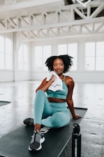 A woman in a teal sports outfit is sitting on a black exercise mat in a bright, spacious gym. She holds a white towel and appears relaxed. The gym features large windows and industrial-style beams across the ceiling.