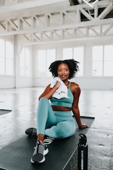 A woman in a teal sports outfit is sitting on a black exercise mat in a bright, spacious gym. She holds a white towel and appears relaxed. The gym features large windows and industrial-style beams across the ceiling.