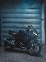 A sleek blue motorcycle is parked in front of a metallic, roll-down shutter. The lighting is low, creating a moody atmosphere, while the bike's sharp lines and modern design are emphasized. There is a touch of industrial and urban feel, with the rough texture of the ground adding to the scene.