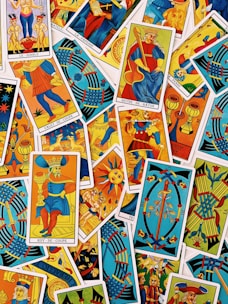A colorful assortment of tarot cards is laid out, featuring a variety of illustrations, including figures, symbols, and mythical imagery. The cards are overlapping, each with vibrant colors and intricate designs, representing different suits and trumps within the tarot deck.