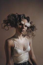 A person with curly hair adorned with feathers, wearing a silky camisole top with lace detailing and standing against a neutral background. The lighting is soft and creates a dramatic shadow effect.