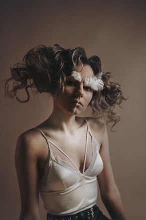 A person with curly hair adorned with feathers, wearing a silky camisole top with lace detailing and standing against a neutral background. The lighting is soft and creates a dramatic shadow effect.