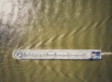 An aerial view of a large cargo ship traveling through greenish-brown water, with visible deck equipment and infrastructure.