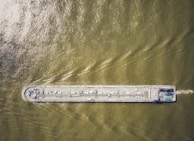 An aerial view of a large cargo ship traveling through greenish-brown water, with visible deck equipment and infrastructure.