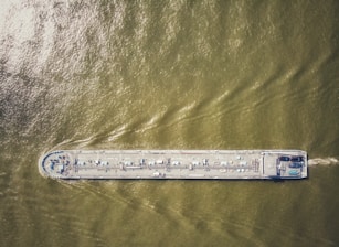 An aerial view of a large cargo ship traveling through greenish-brown water, with visible deck equipment and infrastructure.
