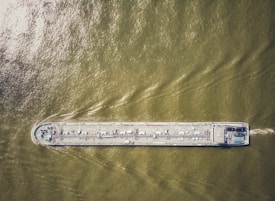 An aerial view of a large cargo ship traveling through greenish-brown water, with visible deck equipment and infrastructure.