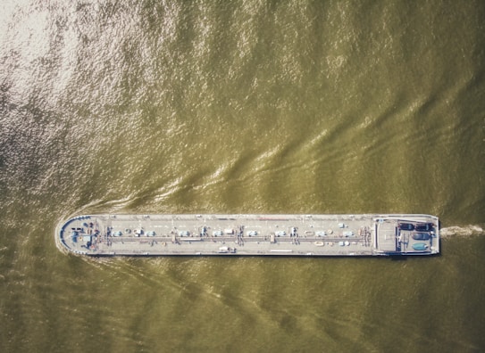 An aerial view of a large cargo ship traveling through greenish-brown water, with visible deck equipment and infrastructure.