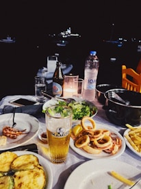 A dining table is set with a variety of seafood dishes, including calamari and grilled vegetables, accompanied by a glass of beer and a bottle of mineral water. The background features a dark night setting with blurred lights from boats.