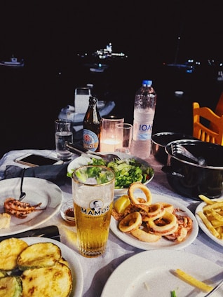 A dining table is set with a variety of seafood dishes, including calamari and grilled vegetables, accompanied by a glass of beer and a bottle of mineral water. The background features a dark night setting with blurred lights from boats.