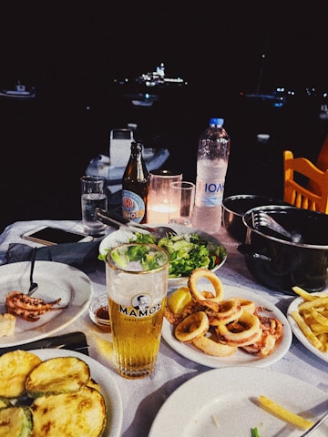 A dining table is set with a variety of seafood dishes, including calamari and grilled vegetables, accompanied by a glass of beer and a bottle of mineral water. The background features a dark night setting with blurred lights from boats.