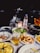 A dining table is set with a variety of seafood dishes, including calamari and grilled vegetables, accompanied by a glass of beer and a bottle of mineral water. The background features a dark night setting with blurred lights from boats.