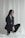 A woman is dressed in a black suit with high-heeled shoes, kneeling on a white fabric backdrop. Her long hair falls on her shoulders, and she gazes slightly away from the camera.