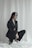 A woman is dressed in a black suit with high-heeled shoes, kneeling on a white fabric backdrop. Her long hair falls on her shoulders, and she gazes slightly away from the camera.